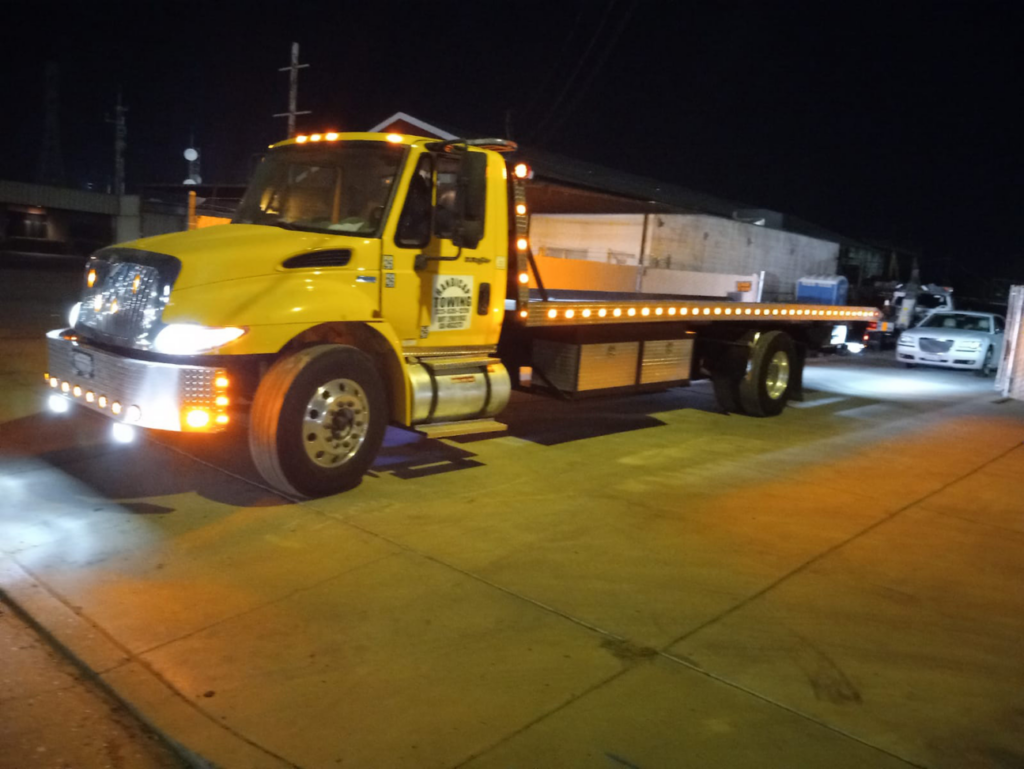 A bright yellow flatbed tow truck with its lights on at night, ready for service from A&A Towing Services in Bakersfield, CA.