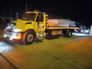 A bright yellow flatbed tow truck with its lights on at night, ready for service from A&A Towing Services in Bakersfield, CA.