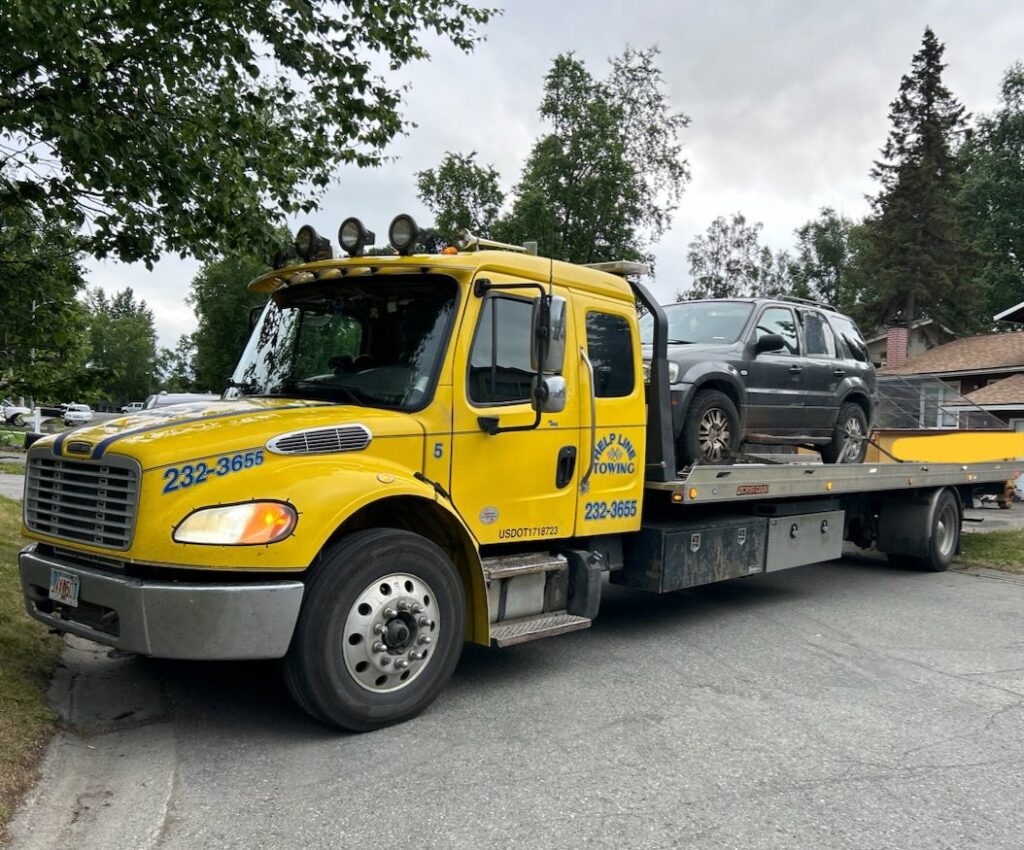 A yellow flatbed tow truck from Help Line Towing transporting a grey SUV on a residential street in Wasilla, AK.