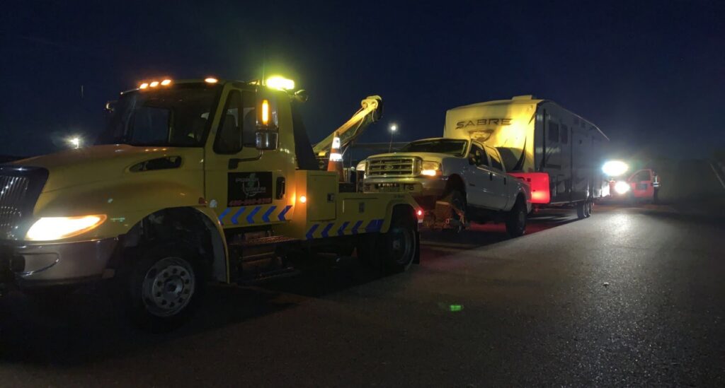 A yellow heavy-duty tow truck from Power Towing & Transport towing a pickup truck and a large travel trailer at night in Phoenix, AZ.