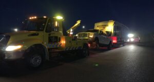 A yellow heavy-duty tow truck from Power Towing & Transport towing a pickup truck and a large travel trailer at night in Phoenix, AZ.