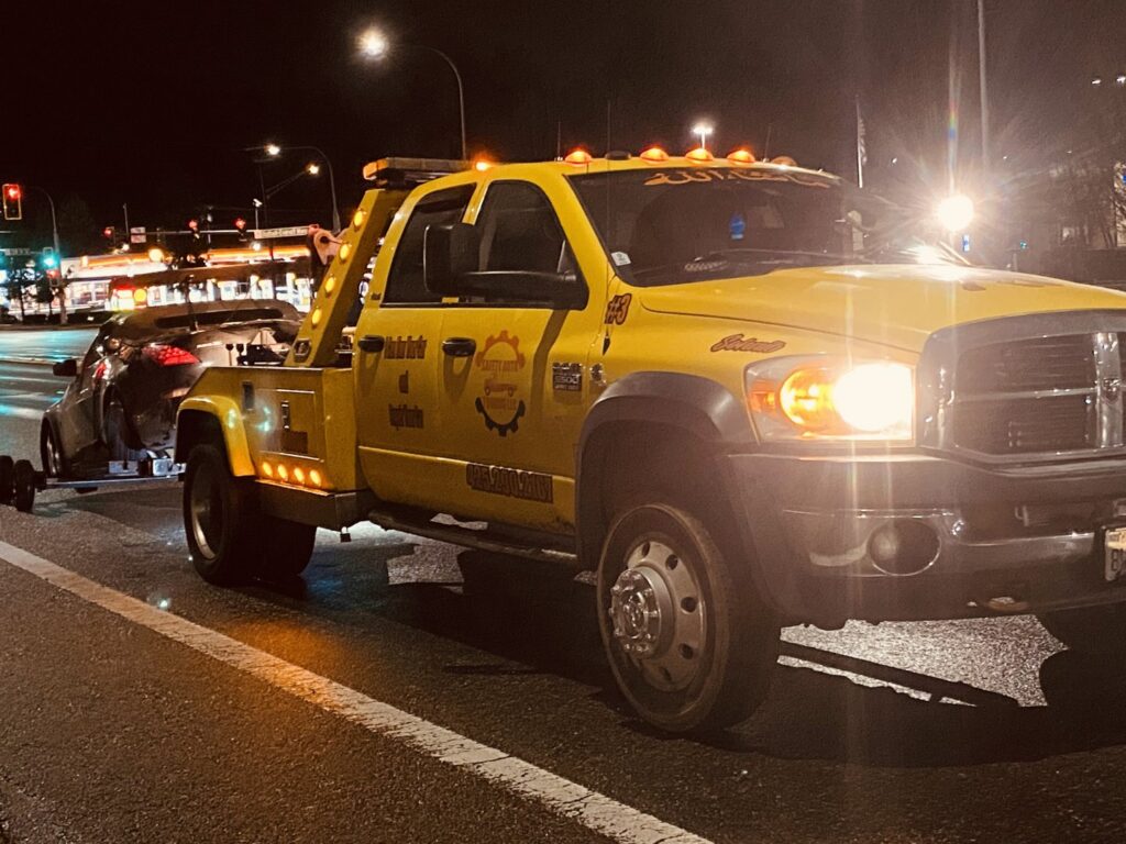 A yellow wrecker tow truck from Safety AUTO Towing LLC in Seattle, WA, towing a silver car at night.