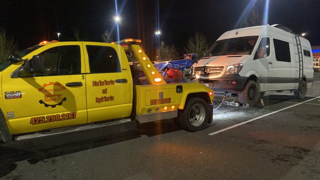A yellow wrecker tow truck from Safety AUTO Towing LLC in Seattle, WA, towing a white van at night.