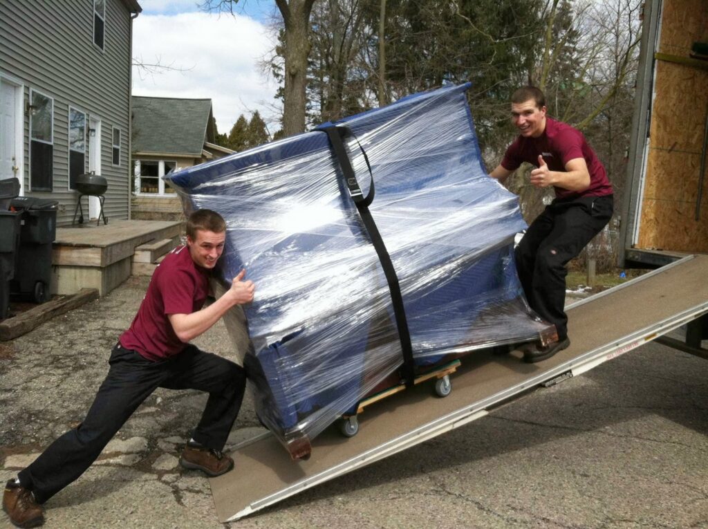 Two movers from Your Friends - Moving Services pushing a wrapped furniture piece up a truck ramp in Lansing, MI.