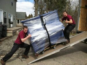 Two movers from Your Friends - Moving Services pushing a wrapped furniture piece up a truck ramp in Lansing, MI.